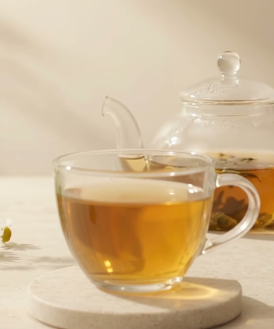 Xícara de vidro com chá dourado do blend SÓMA sobre base de pedra clara, com bule ao fundo